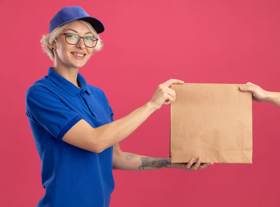 包装身穿蓝色制服 头戴鸭舌帽的年轻女送货员站在粉红色的墙上 友好地微笑着接受纸包裹同时制服朋友