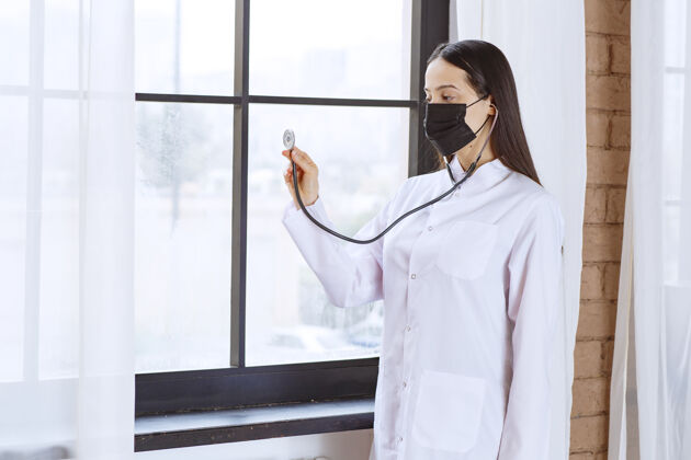 过程戴着黑色口罩的医生站在窗边用听诊器检查心跳从业者姿势制服