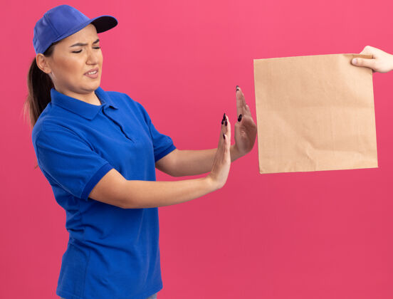 拿着身穿蓝色制服 戴着帽子的年轻送货员站在粉色的墙上拒绝拿纸包裹拒绝送货女人