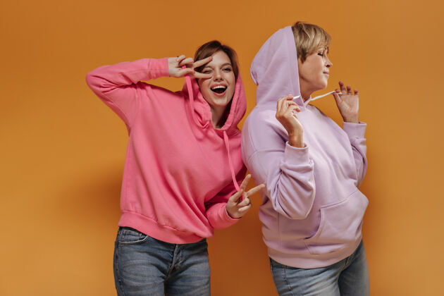 年轻活泼的年轻女孩穿着粉色运动衫 展示着和平的标志 在橙色的背景下和穿着淡紫色凉风连帽衫的现代女性眨眼并合影女成熟粉色