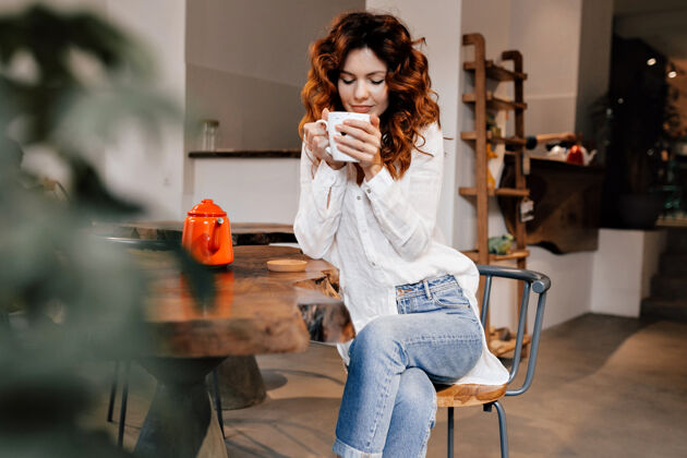 杯子穿着白色衬衫和牛仔裤的时尚可爱的年轻女士坐在舒适的咖啡馆里喝着美味的咖啡 下班后放松夏天女人人