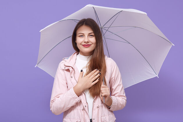 喜悦一位年轻的欧洲妇女 把一把伞孤立在淡紫色的墙上 手放在胸前 真诚地微笑 感激 表达她愉快的态度热情乐趣伞