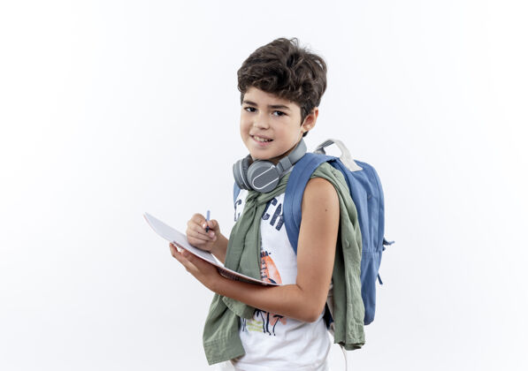 耳机高兴的小男生背着书包 戴着耳机 手里拿着笔记本 笔孤立地放在白色背景上 还有复印空间小的拿着笔记本