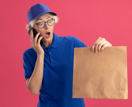 惊喜身穿蓝色制服 头戴鸭舌帽 手拿纸包的年轻送货员在粉色墙壁上讲手机时 显得既惊讶又惊讶帽子惊喜制服