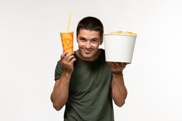 电影正面图身穿绿色t恤的年轻男性手持土豆cips和苏打水在白墙上看电影男性孤独电影年轻男性电影观点