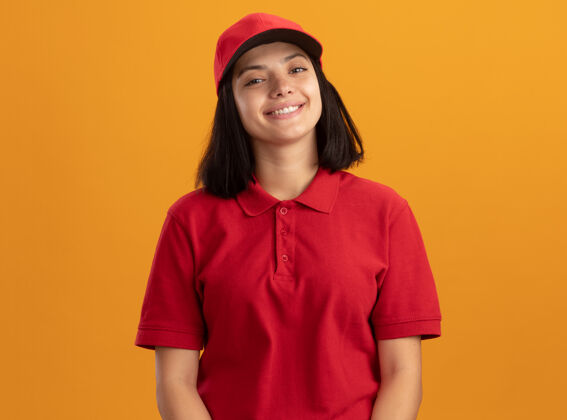 微笑身着红色制服 戴着帽子的快乐的年轻送货女孩站在橙色的墙上 开心地微笑着欢呼年轻站着