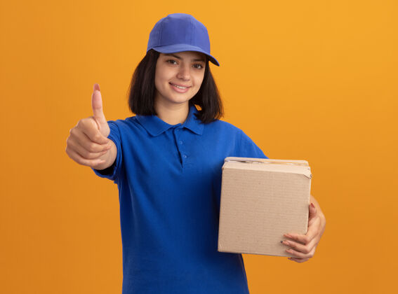 递送身穿蓝色制服 戴着帽子 拿着纸板箱的年轻送货女孩站在橙色的墙上 愉快地微笑着竖起大拇指微笑女孩表演