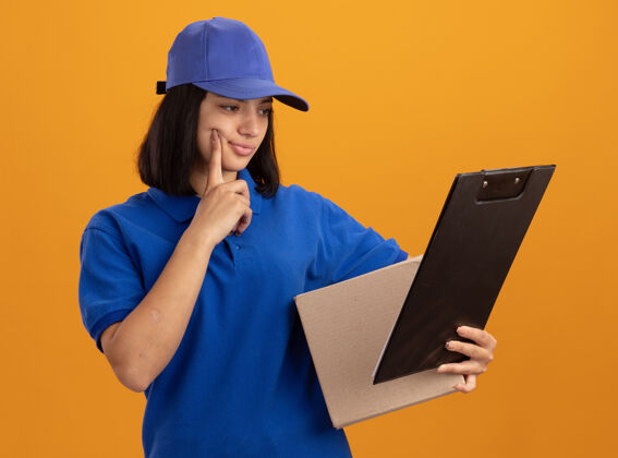 剪贴板身穿蓝色制服 戴着帽子的年轻送货女孩拿着纸板箱和剪贴板 站在橙色的墙上 表情困惑地看着它年轻帽子困惑