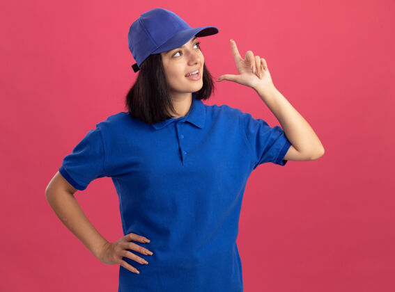 显示身穿蓝色制服 戴着帽子的年轻送货员站在粉红色的墙上 一边愉快地 情绪激动地看着自己的食指年轻情感幸福