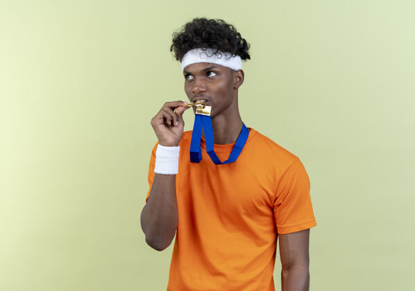 非裔美国人看着身边年轻的美国黑人运动男子戴着头带和腕带举行和咬伤奖牌在脖子上孤立的绿色背景颈部持有咬