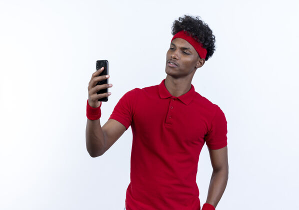 印象令人印象深刻的年轻非裔美国人的运动男子戴着头带和腕带举行 看着电话上的白色背景隔离运动腕带头带