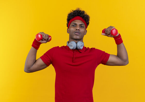耳机自信的年轻美国黑人运动男子戴着头带和腕带 脖子上戴着耳机 举着黄色背景的哑铃穿运动脖子