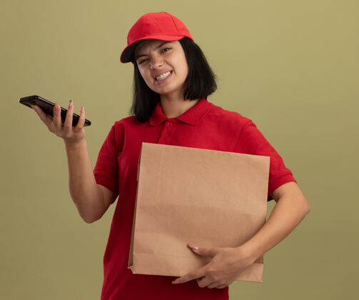 包装穿着红色制服 戴着帽子 拿着纸包的年轻送货女孩站在明亮的墙上 脸上带着恼怒的表情交货立场恼火