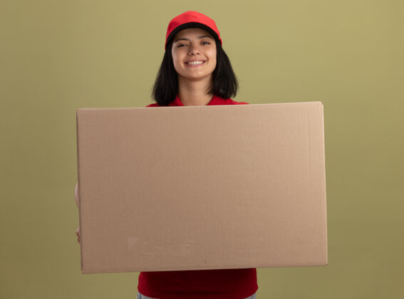 大快乐的年轻送货女孩 穿着红色制服 戴着帽子 手里拿着一个大大的纸箱 脸上带着微笑站在明亮的墙上制服年轻交付