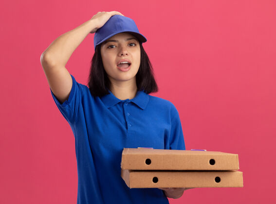披萨身穿蓝色制服的年轻送货员手里拿着比萨饼盒 看上去很困惑 因为她站在粉红色的墙上犯了个错误制服女孩错误