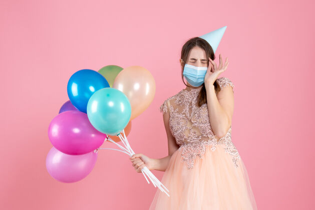 生日正面图可爱的派对女孩戴着派对帽和医疗面罩闭上眼睛拿着五颜六色的气球空间帽子人