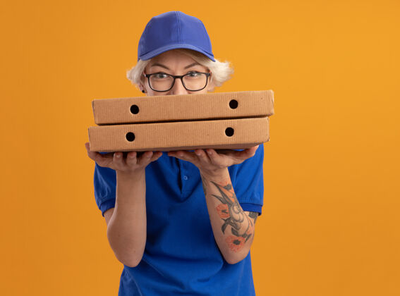 戴着身穿蓝色制服 戴着眼镜 拿着披萨盒的年轻送货员在橙色的墙上狡黠地微笑着年轻人制服盒子