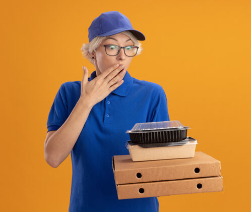 制服身穿蓝色制服 戴着眼镜 拿着比萨饼盒和食品包的年轻送货员惊讶地用手捂住嘴巴 把橘黄色的墙壁翻过来年轻人送货眼镜
