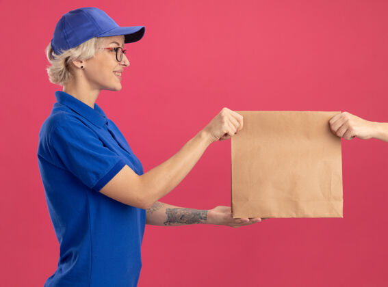 制服身穿蓝色制服 头戴鸭舌帽的年轻女送货员站在粉红色的墙上 友好地微笑着接受纸包裹年轻送货女人