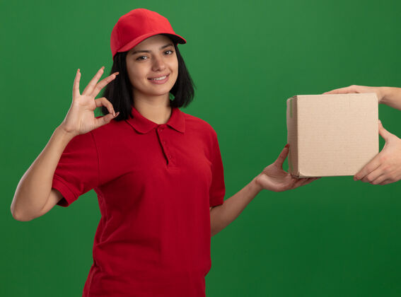顾客身着红色制服 头戴鸭舌帽的年轻送货员向站在绿色墙上的顾客微笑友好地展示ok标志表演女孩站立