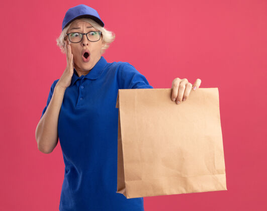 女人穿着蓝色制服 戴着眼镜 拿着纸包裹的年轻送货员看着它 惊讶地看着粉色的墙壁惊人惊喜包装