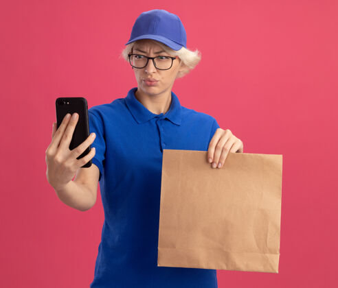 制服穿着蓝色制服 戴着帽子的年轻送货员拿着纸包 看着自己的智能手机屏幕 不高兴地看着粉色的墙壁送货移动女人
