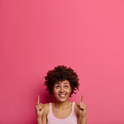 卷发积极的民族女性为您的广告在头顶上方的版面竖起手指 吸引人们的注意力 高高兴兴地微笑 孤零零地站在粉红色的墙上 给出建议 推销产品请年轻积极