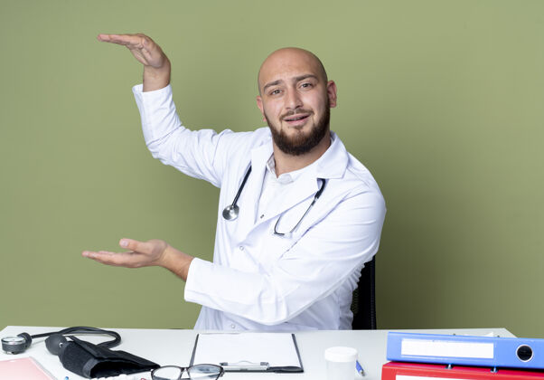 展示高兴的年轻秃头男医生穿着医用长袍和听诊器坐在办公桌上工作 医疗工具显示大小隔离在绿色背景上医疗男性工具