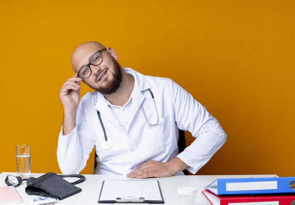 工具年轻的秃顶男医生穿着医用长袍 戴着听诊器 戴着眼镜 坐在办公桌旁 橙色背景上隔离着医疗工具请戴眼镜年轻