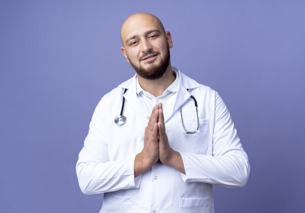 手势高兴的年轻秃头男医生穿着医用长袍和听诊器显示祈祷手势隔离在蓝色背景上穿医疗显示