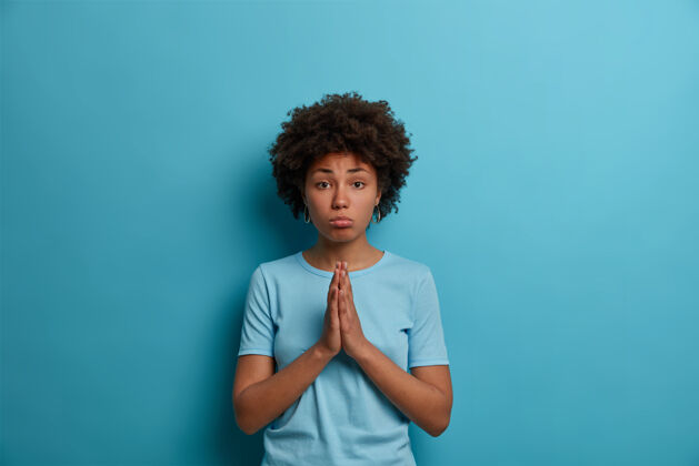 成人愁眉苦脸的非洲头发女人双手合十祈祷 越过蓝色的墙壁恳求 需要你的帮助 乞求帮助 穿着休闲t恤 做出天真的表情随意谢谢手势