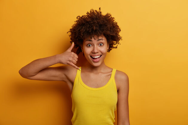 手势友好的积极的女人展示电话标志 想要交谈 与亲密的人保持联系 与人说话 穿着黄色背心 站在室内 给她的电话号码成人肖像休闲