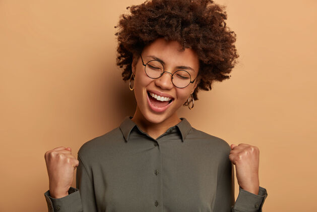 非洲快乐幸运的女商人达成目标 庆祝胜利 与合作伙伴签署盈利协议 戴眼镜和衬衫 隔离在棕色的墙上学生通过了测试积极表情乐观
