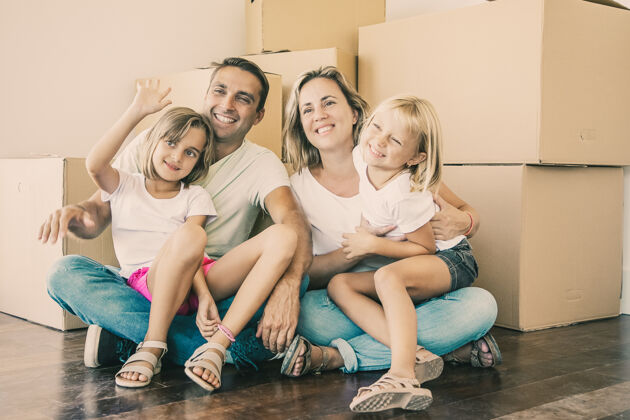 女儿微笑的一家人和孩子们坐在纸板箱旁的地板上 放松着金发女孩站在父亲的腿上挥手致意孩子公寓纸板