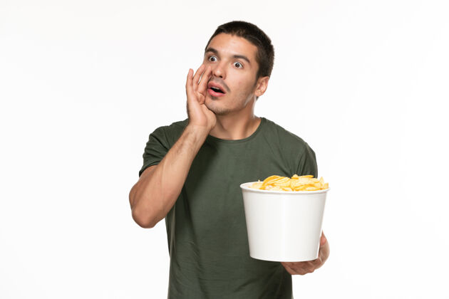 杯子正面图身穿绿色t恤的年轻男性拿着篮子和土豆 在白墙上吃着它们孤独地享受着电影电影电影院视图