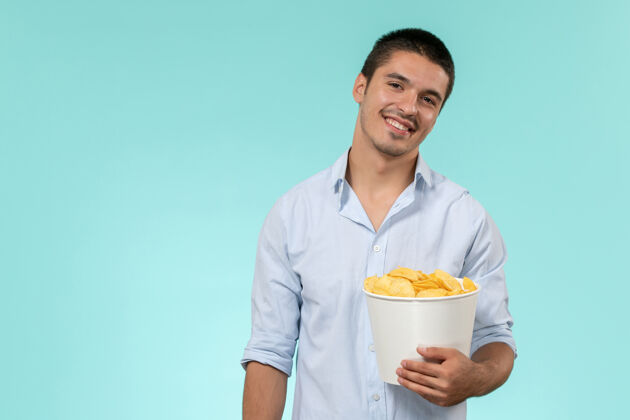 男人正面图：年轻人拿着土豆片 微笑着站在蓝色的墙上 孤独而遥远的男性电影院年轻人土豆视图