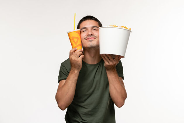 肖像正面图身穿绿色t恤的年轻男性手持土豆cips和苏打水在白墙上看电影男性孤独容器视图成人