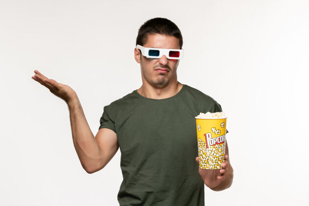 观看正面图身穿绿色t恤的年轻男子戴着d型太阳镜拿着爆米花在白墙上看电影孤独的电影男人肖像视图