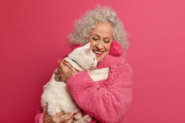 动物美丽的老年白发女子手牵白猫 用爱心呵护自己的宠物 穿着随意 隔着粉红色的墙 闲暇时间在一起 检疫期间呆在家里放松猫朋友