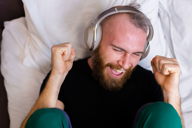 家伙快乐的白人男子戴着耳机躺在床上听他最喜欢的音乐 独自休息 跳舞无线房子科技