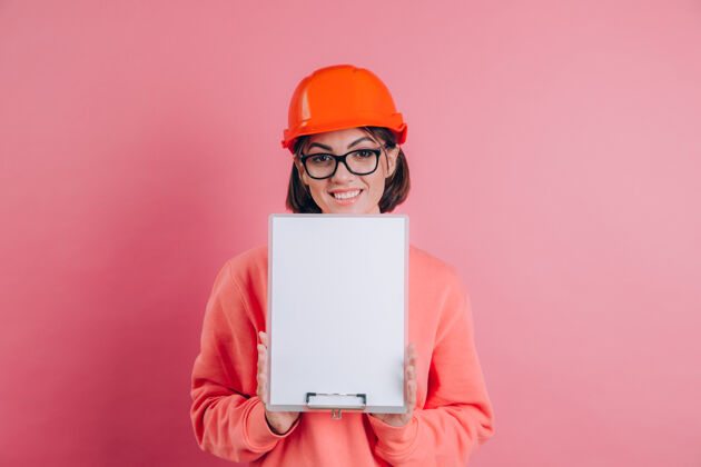 承包商面带微笑的女建筑工人手持白色的空白标志牌 背景是粉色的建筑头盔剪贴板一个帽子