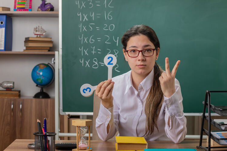 手指年轻的女老师戴着眼镜 拿着号码牌 讲解着第二课 手指自信地坐在教室黑板前的课桌旁女人拿着书桌