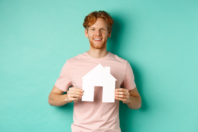 红发房地产概念年轻人红头发 穿着t恤 展示纸房子剪贴画和微笑的幸福 站在薄荷背景复制空间男性房地产房子