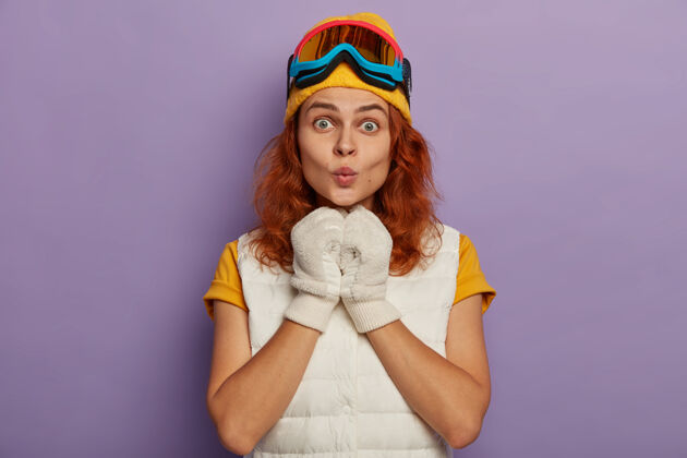 护目镜迷人的红发女人戴着暖和的帽子 滑雪镜和白色套头衫满意脸颊衣服
