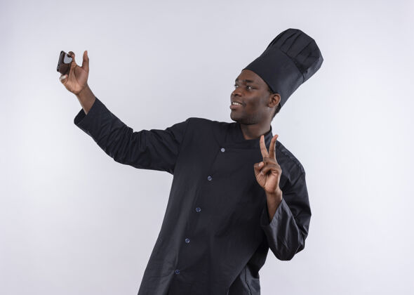 年轻身着厨师制服的年轻的非洲裔美国厨师微笑着看着电话 手势胜利的标志在白色的空白处复制厨师电话