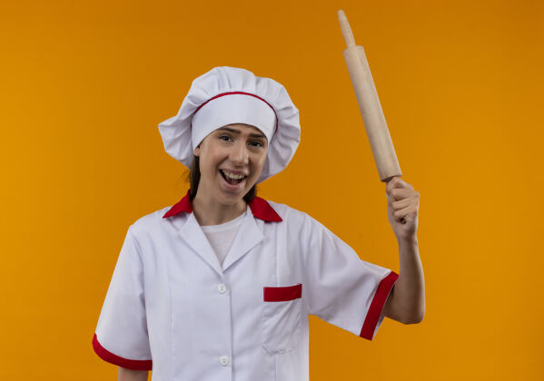 橙色穿着厨师制服的年轻恼怒的白人厨师女孩拿着擀面杖 看着橘子色的相机 还有复印空间复制恼怒制服