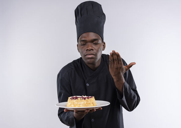 空间身着厨师制服的年轻美籍黑人厨师手持并假装闻到白色盘子上的蛋糕 留有复印空间年轻盘子厨师
