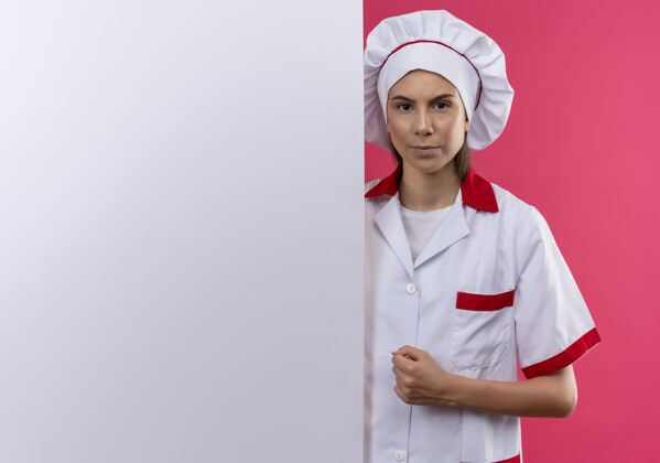 烹饪穿着厨师制服的年轻严肃的白人厨师女孩站在粉色的白色墙壁后面 留有复印空间年轻空间复制