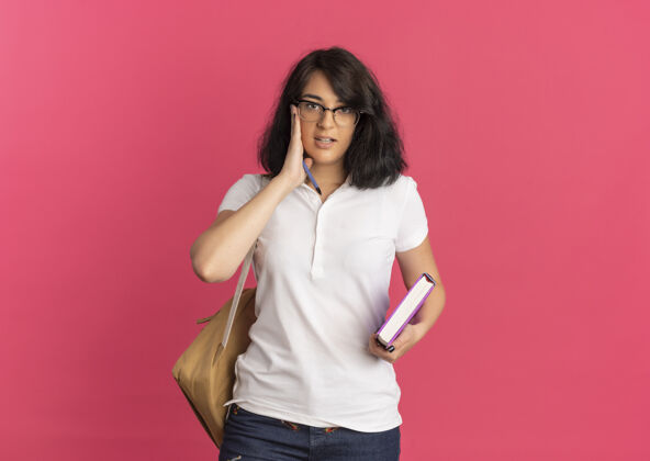 背年轻自信漂亮的高加索女学生戴着眼镜 背着书包 手放在脸上 手里拿着笔和书 粉色的和复印空间眼镜粉色自信