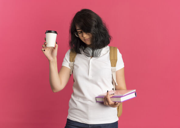 年轻年轻疲惫漂亮的白人女学生 戴着耳机 戴着眼镜 背着书包 拿着咖啡杯和书 闭着眼睛 粉红色的 有复印空间耳机持有包